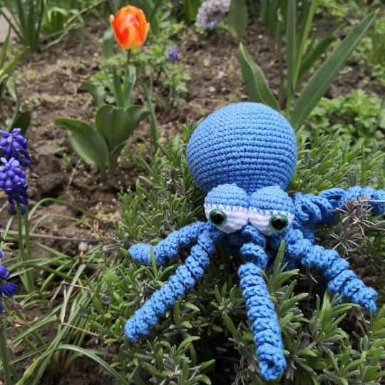 Blue crocheted octopus sitting in a garden setting.